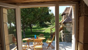 Eine offene Terrasse mit Tisch und Stühlen, Blick auf einen Garten und ein Steinhaus im Hintergrund.