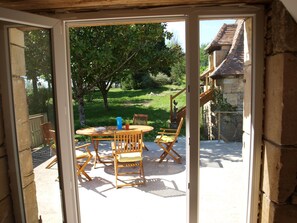 An open sliding glass door leading to a patio with a table, chairs, and a view of a stone building and greenery. - L'hirondelle - la cipière gîtes (Peyzac-le-Moustier)