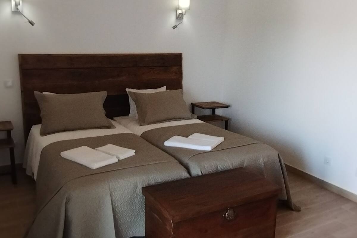 A double bed with two pillows and two folded towels, a wooden nightstand, a small table, and a wooden chest of drawers.