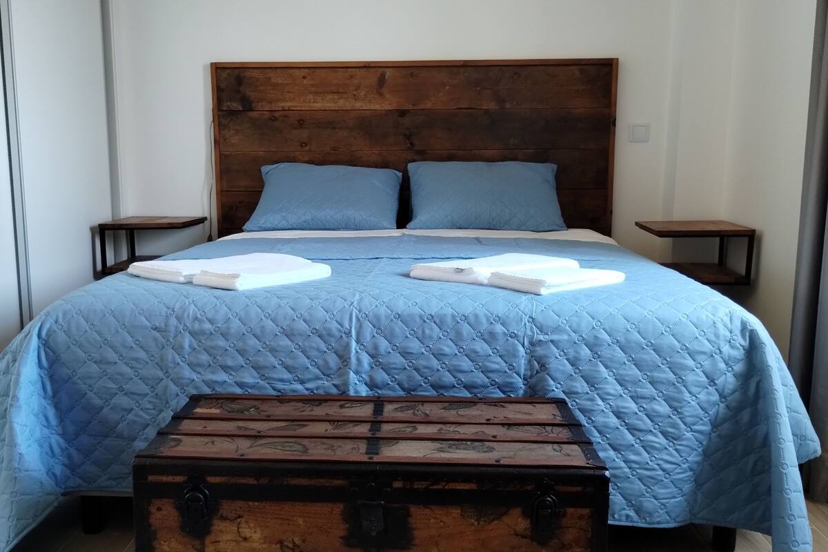 A bed with a blue quilt, two pillows, and two folded towels on it. There is a wooden bench in front of the bed.