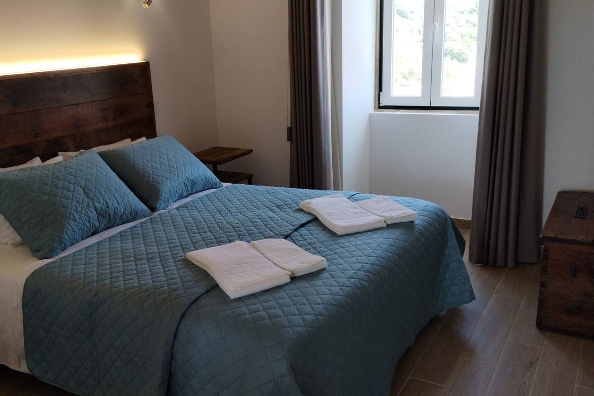 A bedroom with a large bed, two pillows, a bedside table, and a window with curtains.