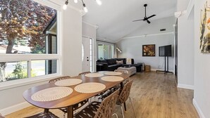A spacious dining area with a wooden table and chairs, a ceiling fan, and a large window with a view of trees. - 3BR · Pool · Hot Tub · BBQ · Garage · Roseville (Roseville)