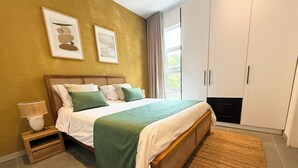 Une chambre à coucher comprenant un lit, une table de chevet, une lampe, des œuvres d’art, une armoire et une vue sur la verdure à l’extérieur.