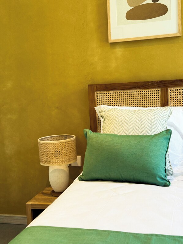 Un lit bien fait, avec un oreiller vert et un oreiller blanc, une table de chevet en bois surmontée d’une lampe en osier, et un tableau encadré accroché au mur.