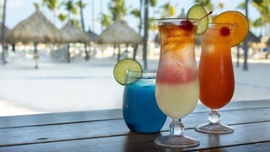 Twee tropische cocktails op een houten tafel, met een strand en palmbomen op de achtergrond.