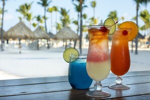 Zwei tropische Cocktails auf einem Holztisch, im Hintergrund Strand und Palmen.