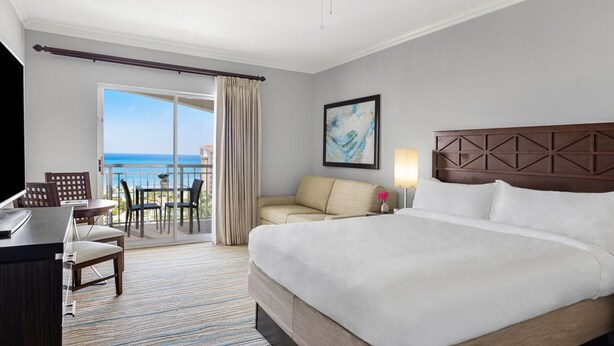 Een hotelkamer met een groot bed, een bureau met stoel, een bank, een balkon met uitzicht op zee, en een schilderij aan de muur.