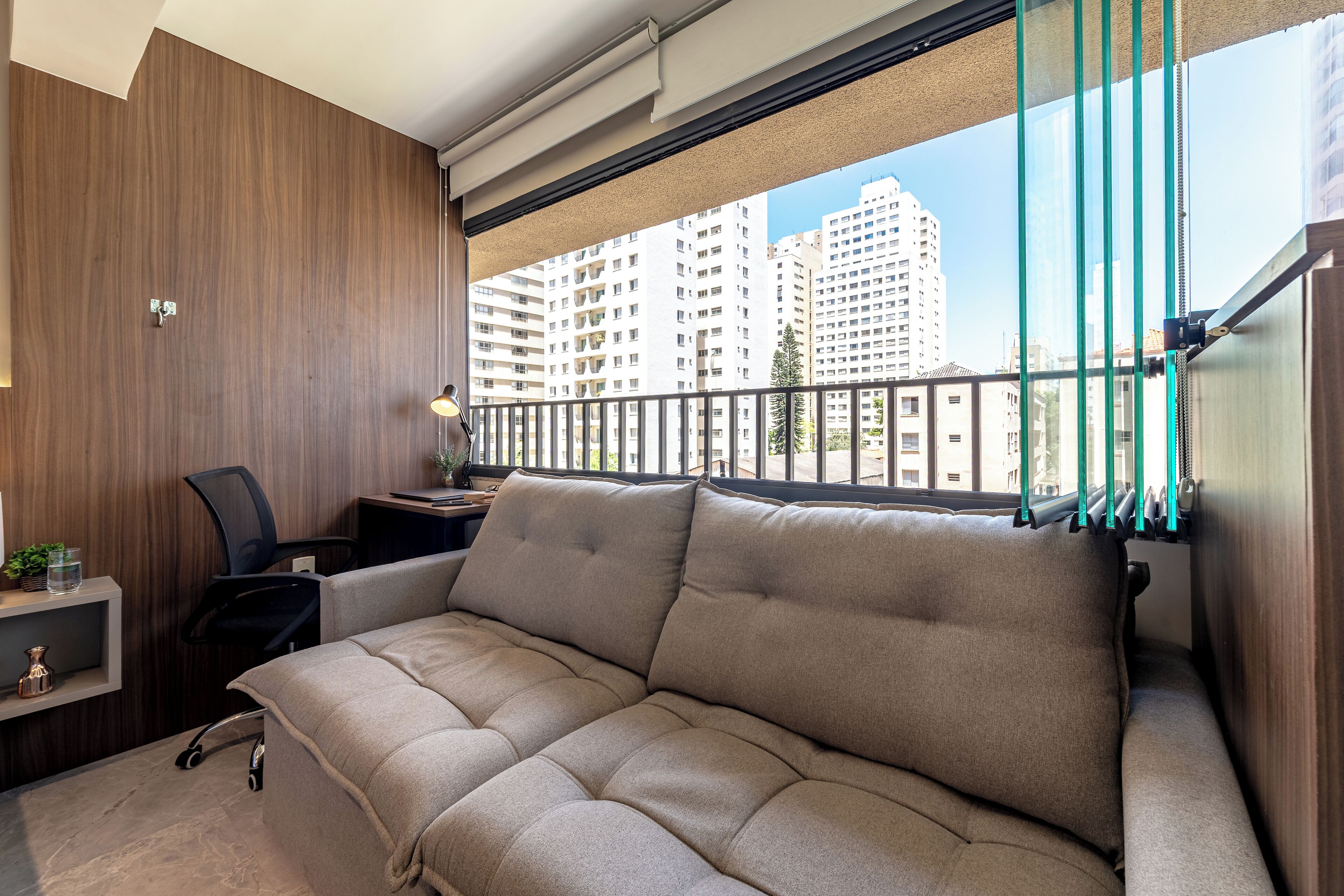 A modern living room with a sofa, a desk, and a chair. There is a window with a view of tall buildings.