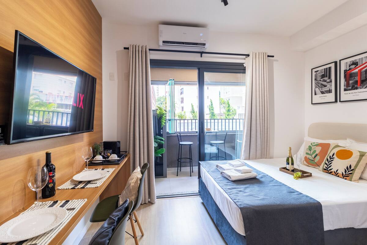 Ein Hotelzimmer mit Bett, Fernseher, Essecke mit Tisch und Stühlen sowie einem Balkon mit Blick auf Gebäude.
