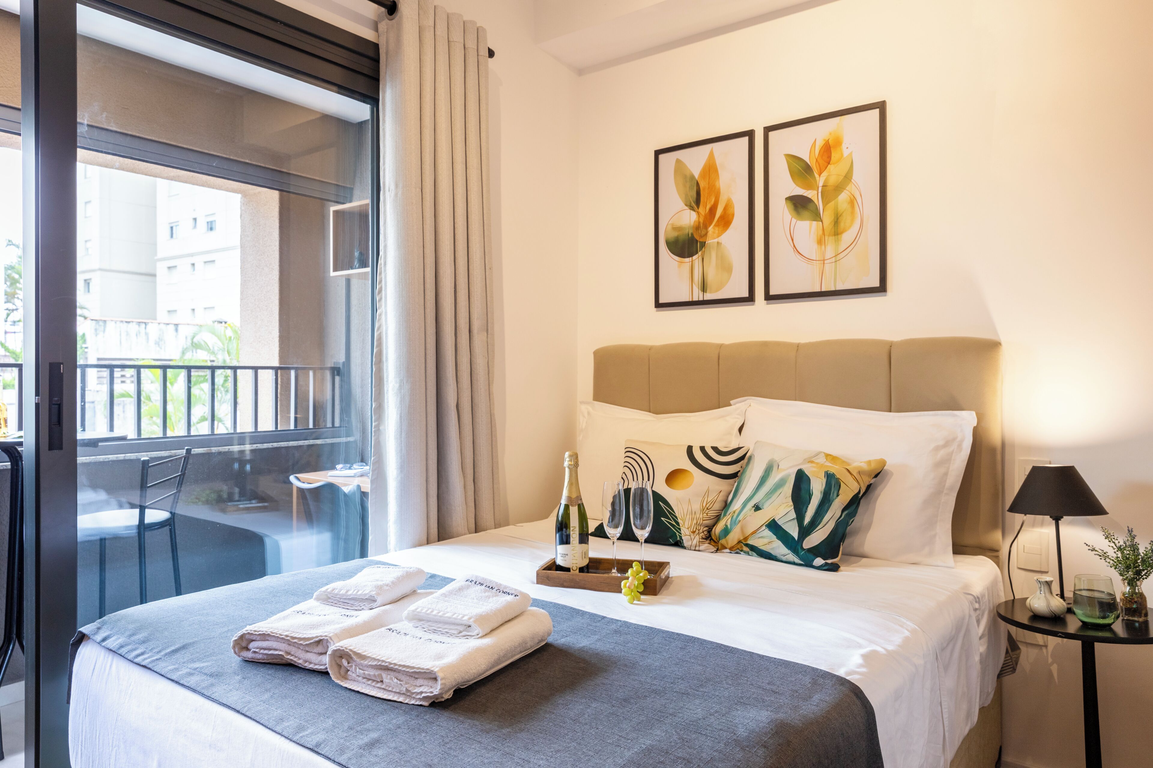 A hotel room with a bed, a balcony, a table with a chair, and two framed floral artworks on the wall.