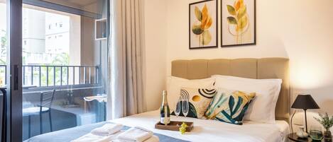 A hotel room with a bed, a balcony, a table with a chair, and two framed floral artworks on the wall.