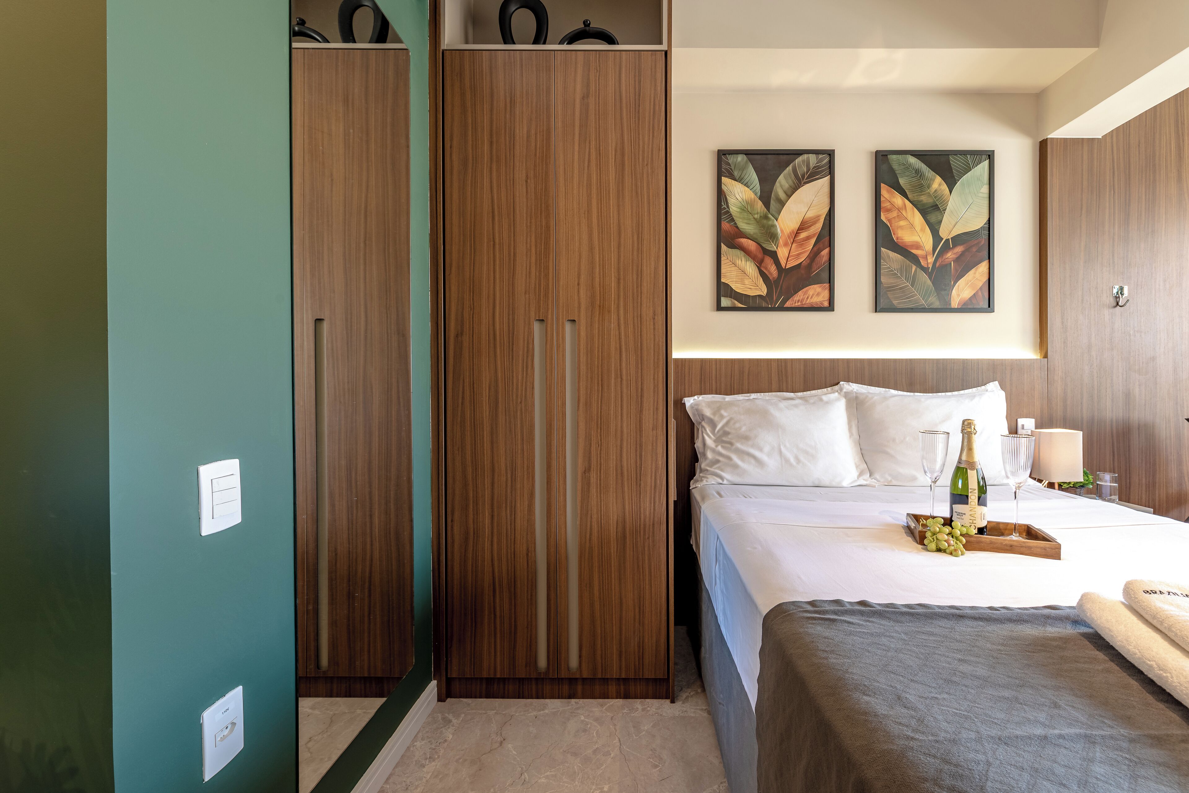 A hotel room with a bed, a wooden wardrobe, and two framed leaf paintings on the wall.