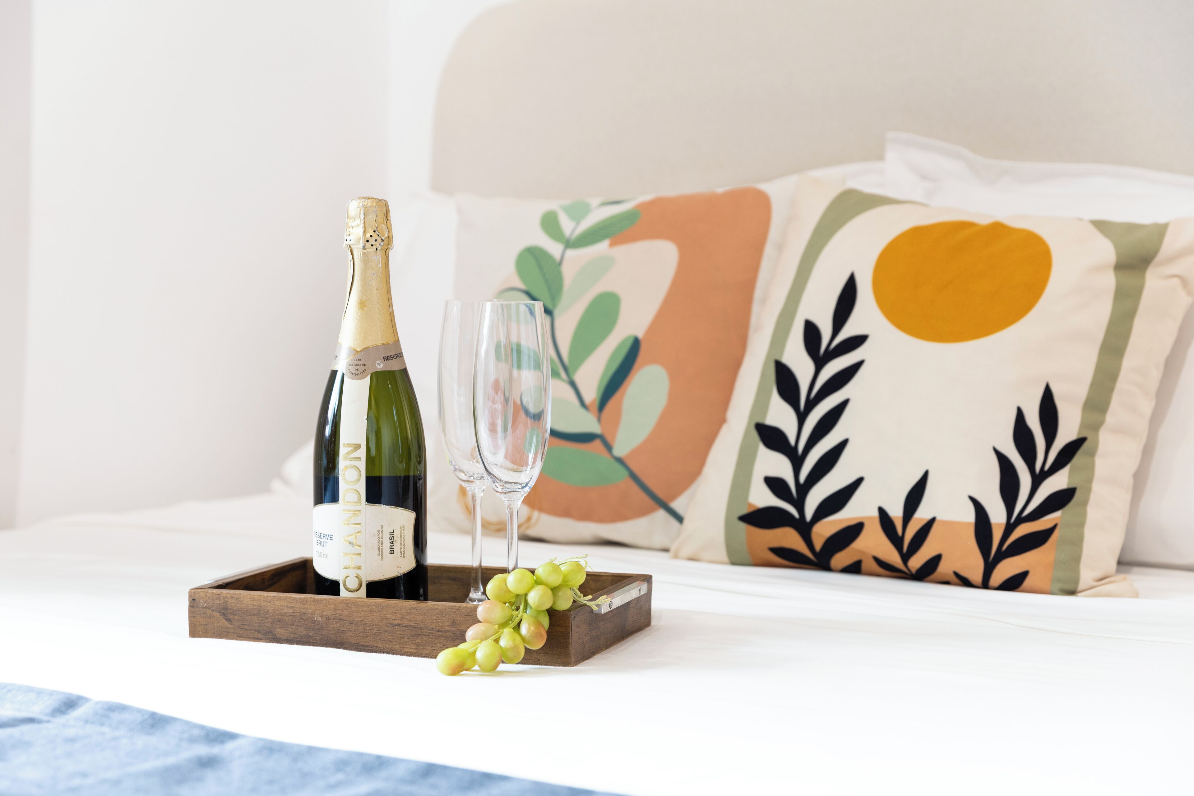 A bed with a wooden tray holding a bottle of champagne, a glass, and grapes.