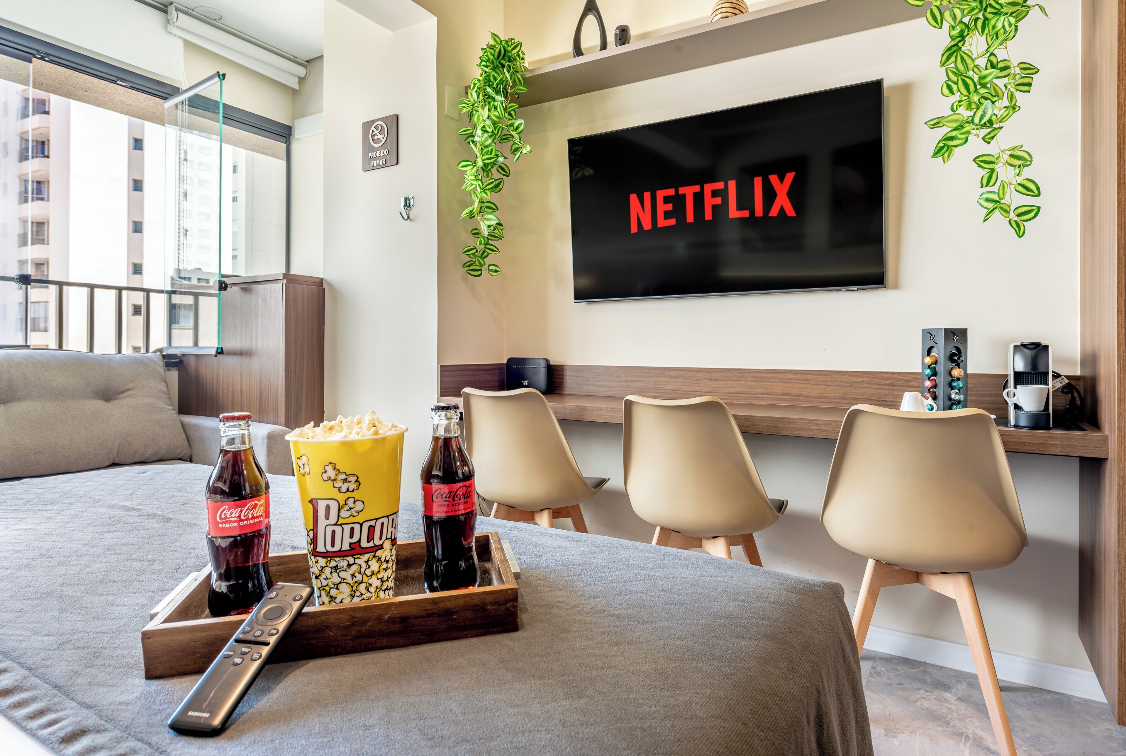 A hotel room with a bed, a TV mounted on the wall, a tray with popcorn and Coca-Cola bottles, and a window with a view of buildings.