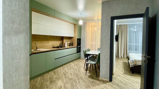 A modern kitchen with light green cabinets, a dining area with a table and chairs, and a bedroom with a bed and curtains. - River View (Raionul Soroca)