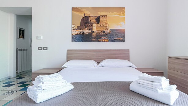 Habitación de hotel con una cama, una pila de toallas ordenadas, cabecero de madera y un cuadro colgado en la pared que representa un castillo costero.