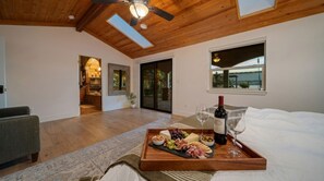 A bedroom with a wooden ceiling, a bed, a breakfast tray with cheese, fruits, nuts, and wine, and a view of a garden through a sliding door.