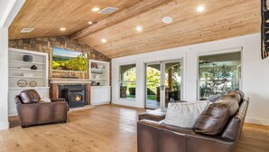 A spacious living room with hardwood floors, a stone fireplace, leather sofas, and large windows offering a view of the outdoors.