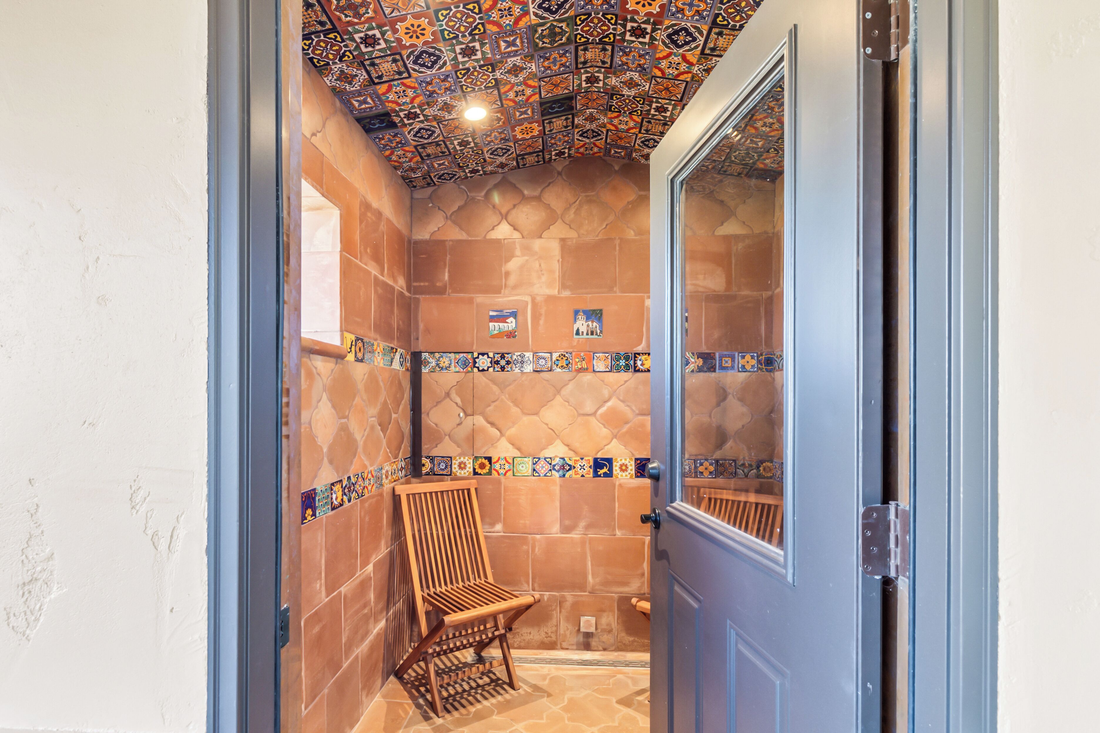 A bathroom with a tiled shower wall, a wooden chair, and a door with a glass panel.