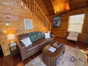 Ein gemütliches Blockhaushaus-Interieur mit einer Holztreppe, einem braunen Ledersofa, einem Holztisch und einem Sessel.