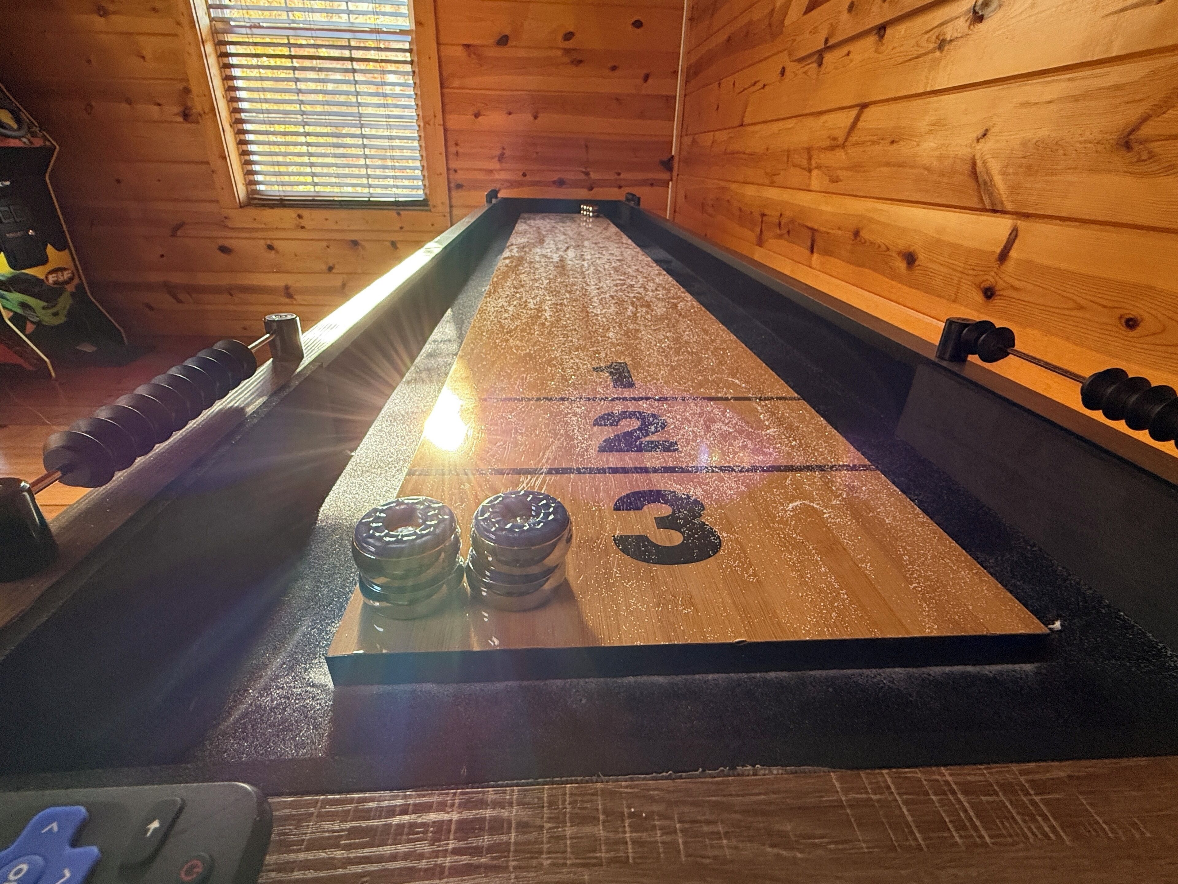 Pöytä, jossa pelataan shuffleboardia, ja jossa on numeroituja pistealueita. Pöydän ympärillä on puuseinät ja yksi ikkuna.