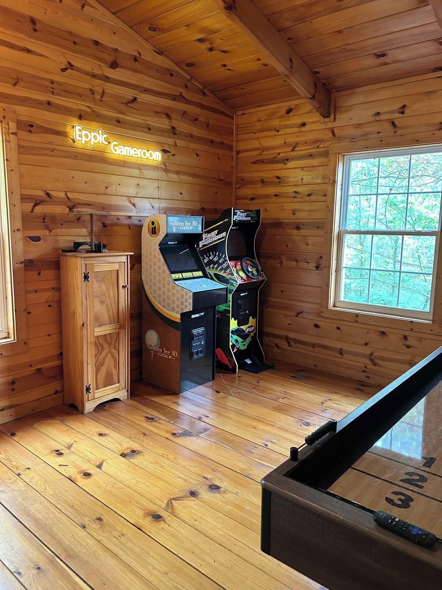 A wooden cabin with a neon sign reading "Epic Gameroom," a wooden cabinet, and two gaming consoles.
