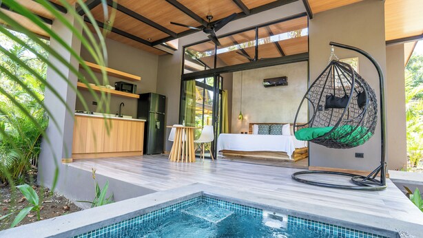Zona de piscina exterior moderna amb una cadira penjant, un llit i una cuina.