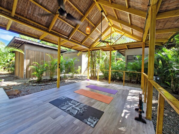 Un espace extérieur abrité, doté de poutres en bois et d’un toit de chaume, comprenant des tapis de sol pour les exercices et un petit élément décoratif aquatique.
