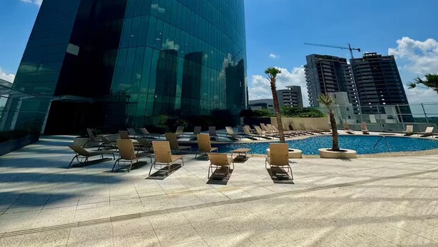 A modern building with a glass facade, a pool area with sun loungers, and a clear blue sky.