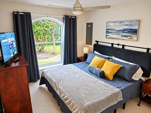 A bedroom with a bed, a TV, a window with curtains, a painting, and a lamp. - Pelican’s Point – Weeki Wachee Main River Home with Gulf Access (Weeki Wachee)