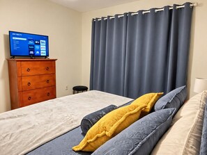 A bedroom with a bed, a wooden dresser, a flat-screen TV, and a large window with curtains. - Pelican’s Point – Weeki Wachee Main River Home with Gulf Access (Weeki Wachee)