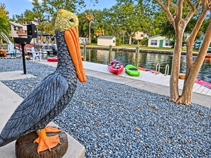 A large bird sculpture on a stone pedestal near a waterfront with kayaks and a dock. - Pelican’s Point – Weeki Wachee Main River Home with Gulf Access (Weeki Wachee)