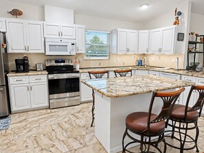 A kitchen with white cabinets, a granite countertop, stainless steel appliances, and a bar stool area. - Pelican’s Point – Weeki Wachee Main River Home with Gulf Access (Weeki Wachee)