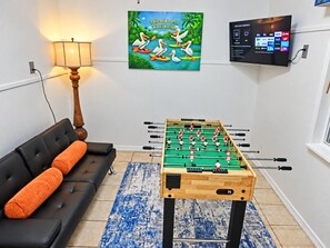 A room with a foosball table, a sofa with orange cushions, a lamp, a TV displaying a channel guide, and a wall art piece. - Pelican’s Point – Weeki Wachee Main River Home with Gulf Access (Weeki Wachee)
