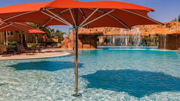 Una piscina con agua cristalina azul, rodeada de sombrillas rojas y una cascada.