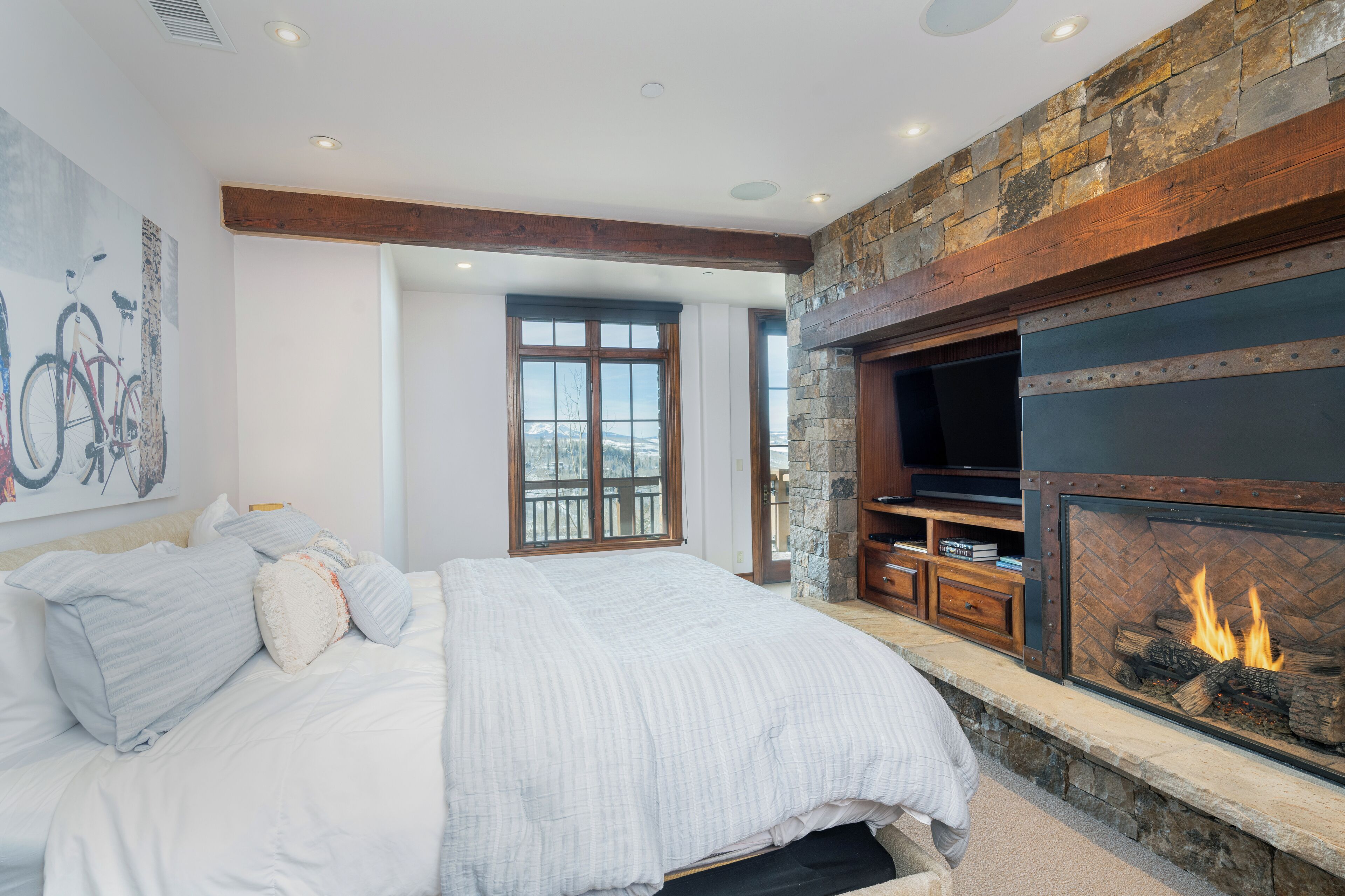 A bedroom with a large bed, a stone fireplace, and a television mounted on the wall.