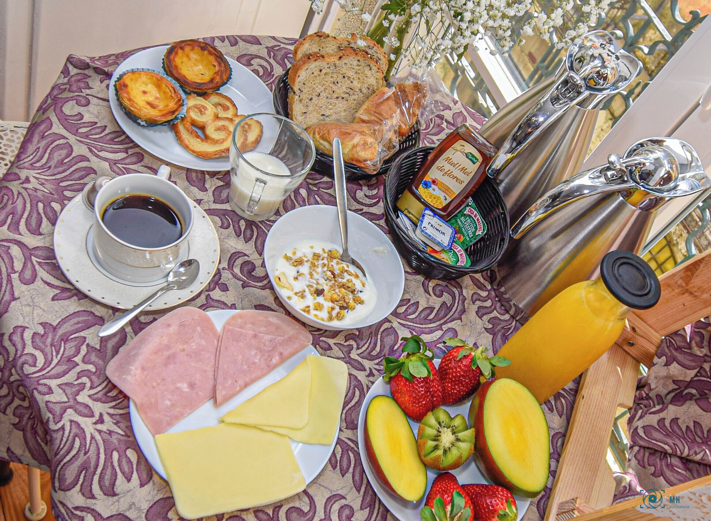 A breakfast spread with bread, pastries, cheese, fruits, and beverages on a patterned tablecloth.