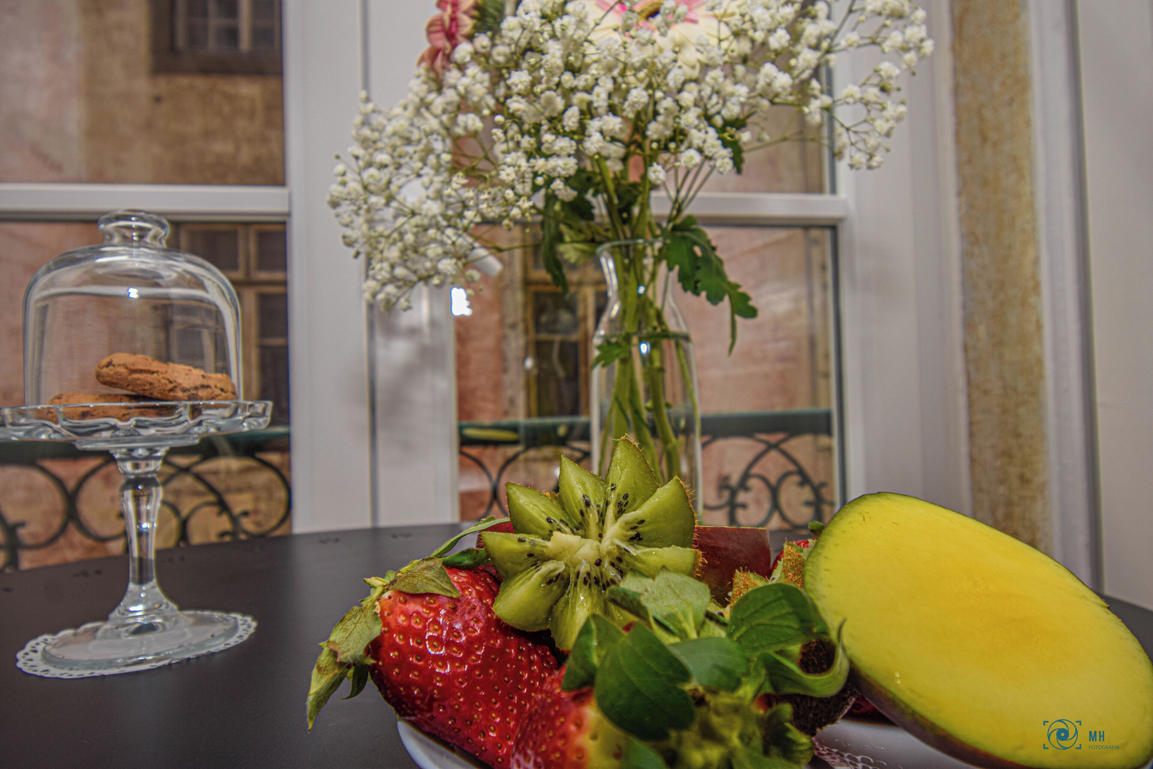 A table with a glass cake stand, a vase with flowers, and a plate with fruit.