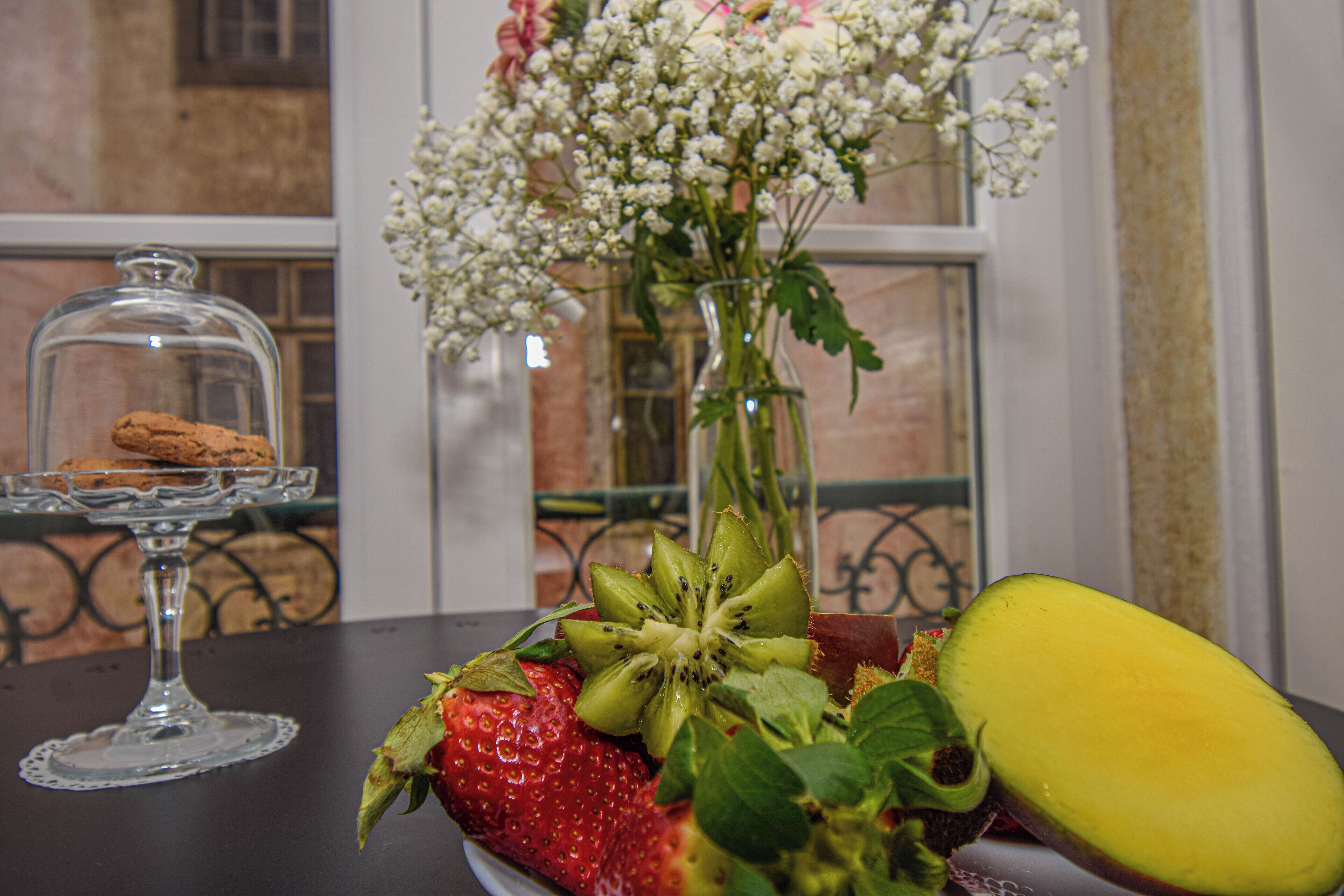 Een tafel met een glazen taartplateau, een vaas met bloemen, en een bord met aardbeien, kiwi en meloen.