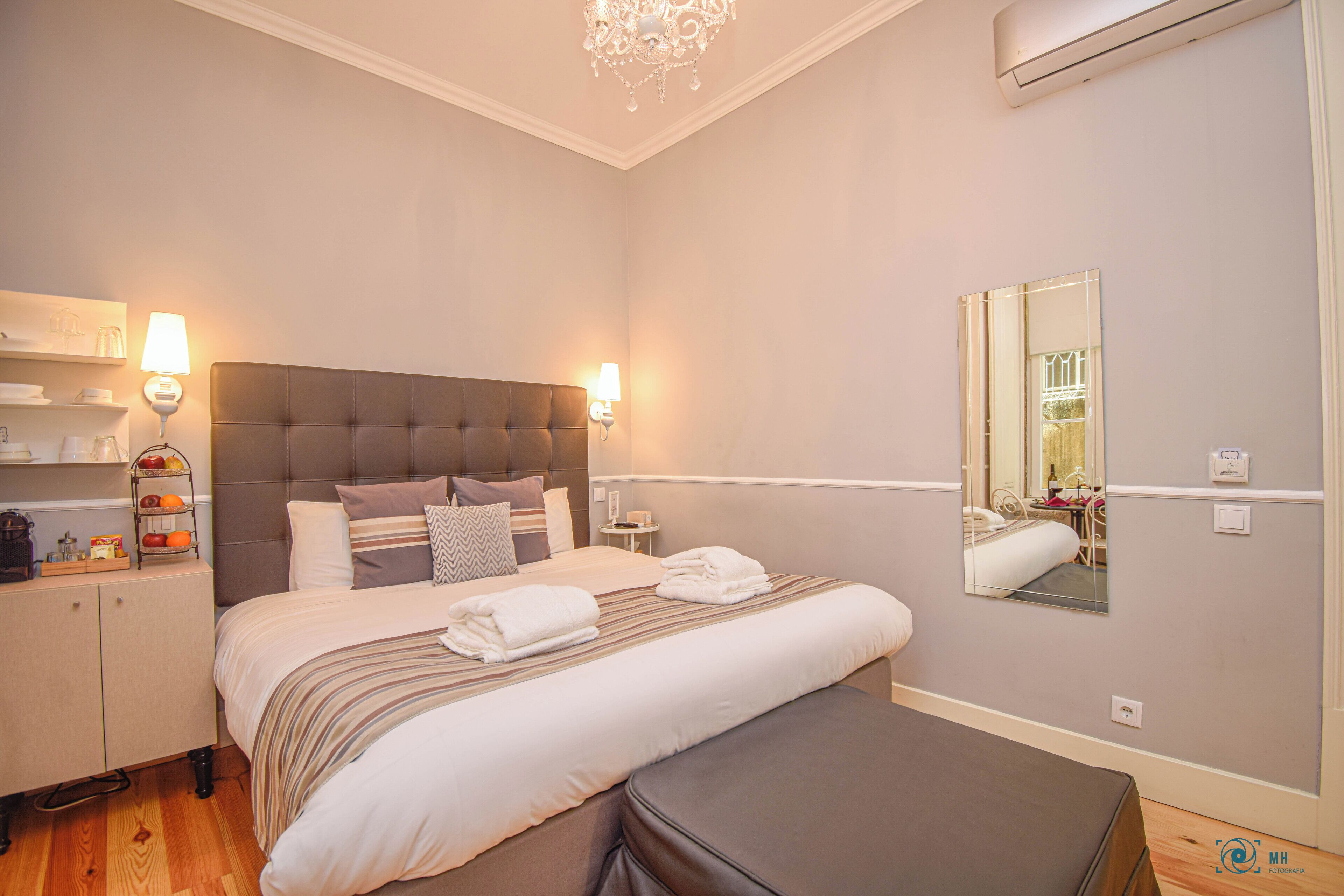 A hotel room with a large bed, bedside tables, a wardrobe, a bench, a chandelier, and a mirror on the wall.