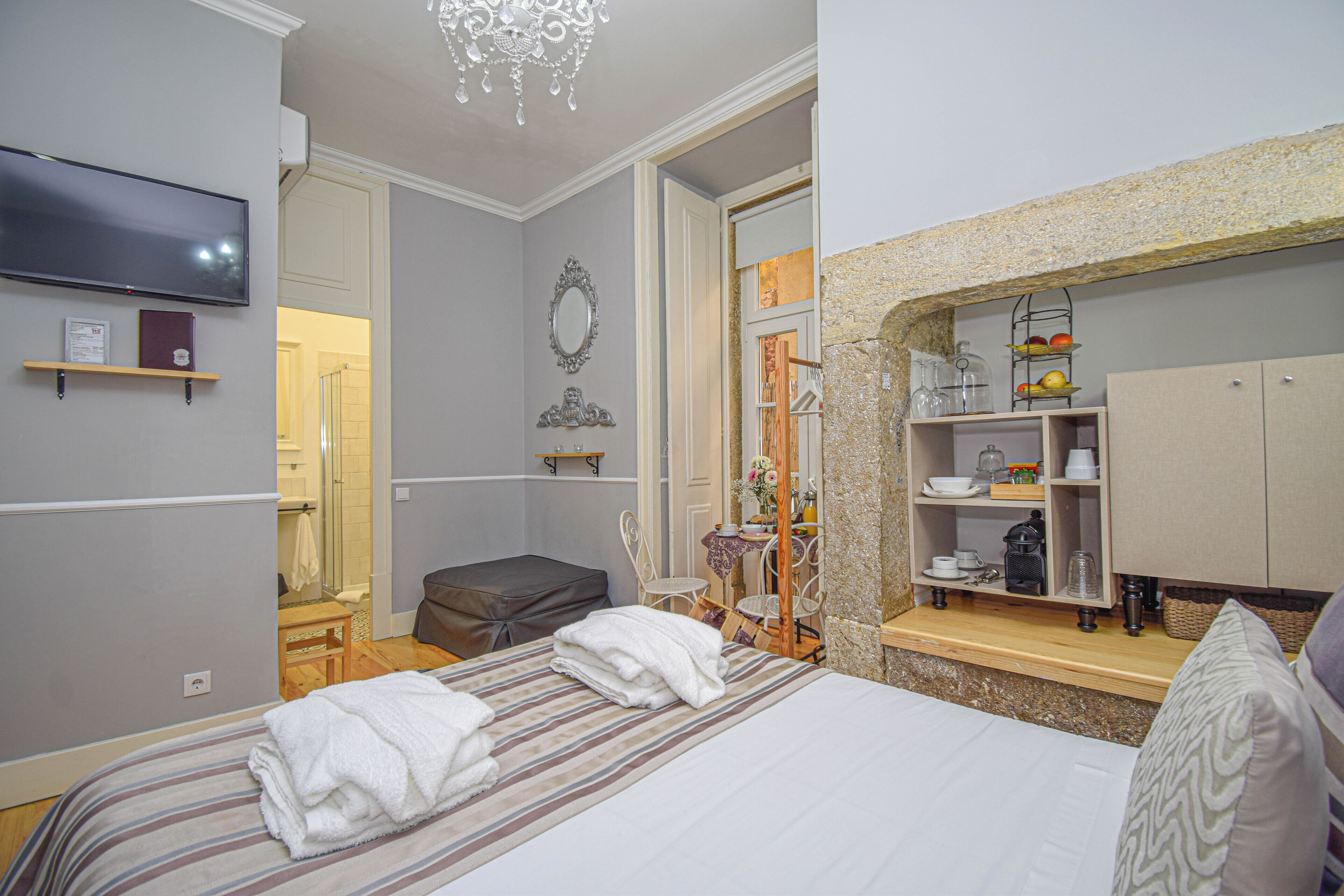 A hotel room with a bed, a TV, a mirror, a small table, and a shelf with kitchenware.