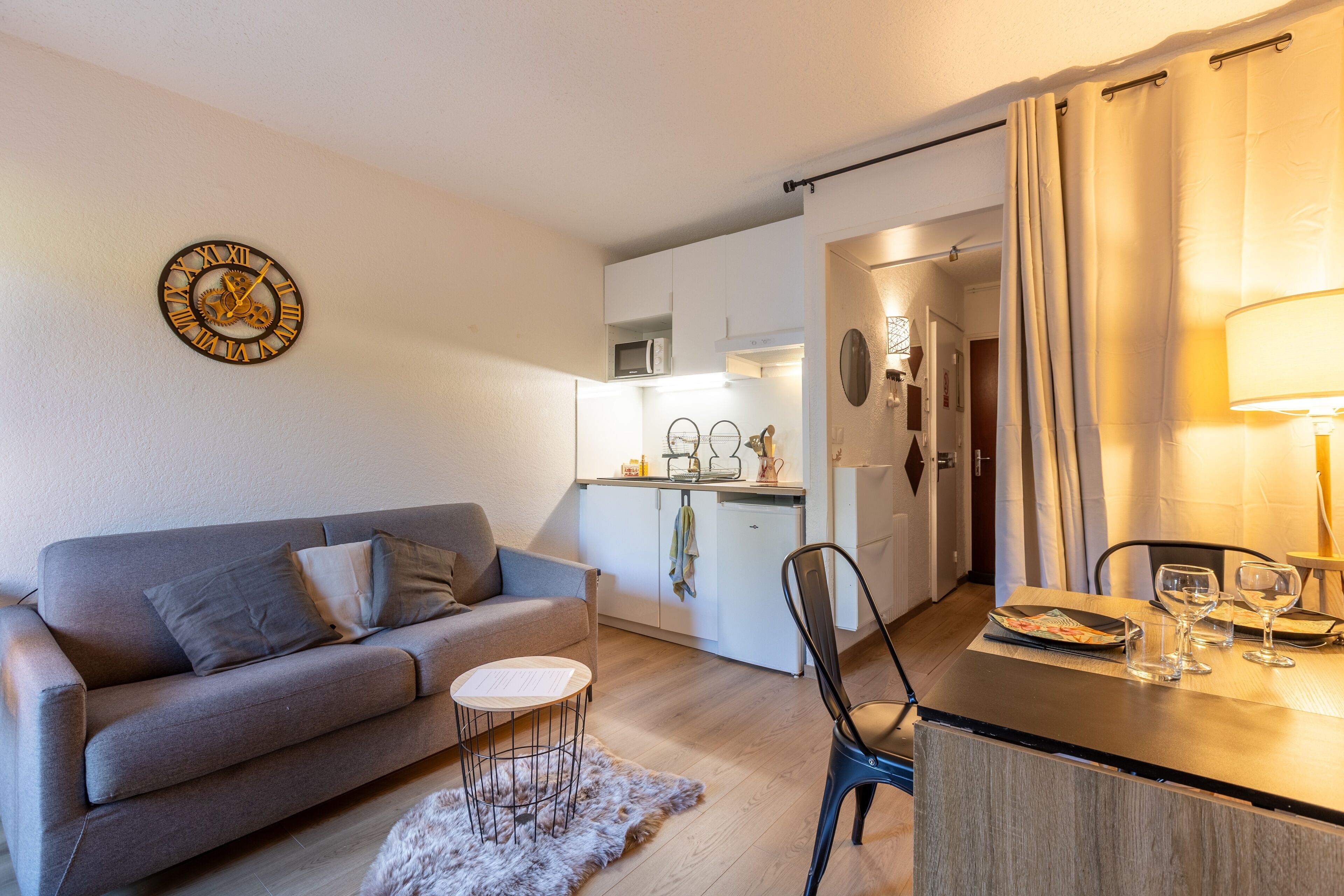 A modern living room with a sofa, a small round table, and a dining area with a chair and wine glasses.