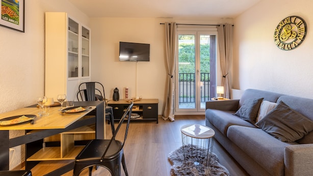 A living room with a sofa, a TV mounted on the wall, a dining table with chairs, and a window with curtains. - Le Cocon Douillet - Proche Télécabines (Les Angles)