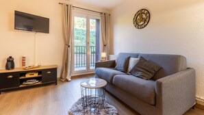 A living room with a grey sofa, a flat-screen TV mounted on the wall, a small round coffee table, a wooden cabinet, and a large window with curtains. - Le Cocon Douillet - Proche Télécabines (Les Angles)
