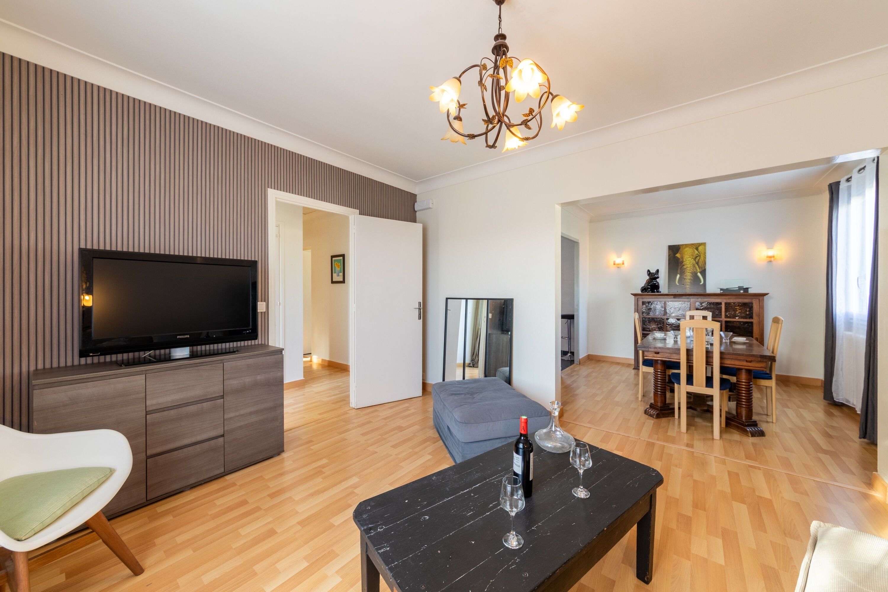 A modern living room with a flat-screen TV, wooden flooring, a dining area with a table and chairs, and a chandelier.