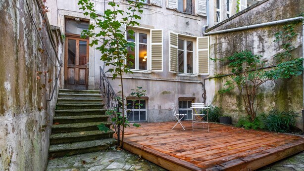 Ein Innenhof mit Holzterrasse, Steinstufen und einem Gebäude mit Fenstern und Fensterläden.