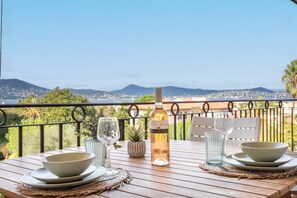 A dining table set with bowls, a wine bottle, and glasses, overlooking a scenic view of hills and a beach. - La Vue Tropézienne - Appartement Climatisé (Saint-Tropez)