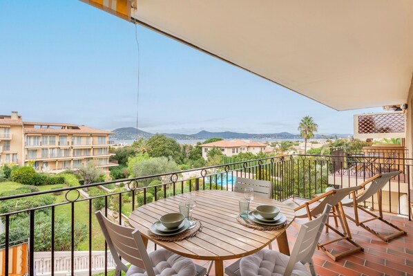 A balcony with a wooden table set for two, overlooking a pool and a cityscape. - La Vue Tropézienne - Appartement Climatisé (Saint-Tropez)