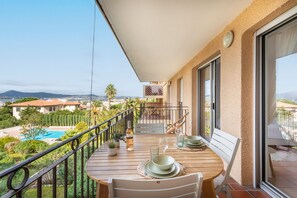 A balcony with a wooden table set for two, overlooking a swimming pool and a mountainous landscape. - La Vue Tropézienne - Appartement Climatisé (Saint-Tropez)