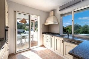 A modern kitchen with a sliding glass door leading to an outdoor patio, built-in cabinets, a stainless steel sink, and a large window offering a view of the outdoors. - Villa Valle Pace Maria - Piscine Privée (Afa)